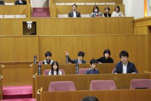 こども議会体験会でこども（1人目）が議員席で手を挙げている写真